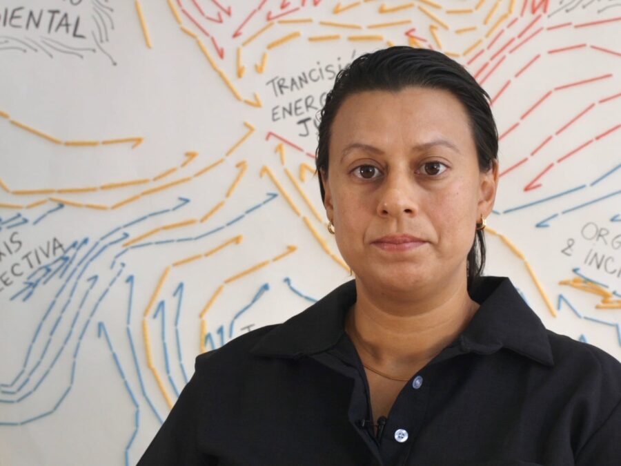 A person with tightly pulled back hair poses in front of a wall filled with writing and blue and orange arrows