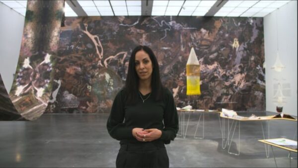 A woman with dark hair dressed in black stands in a gallery with a large mural behind her