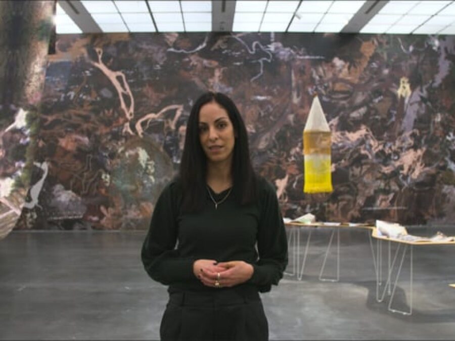 A woman with dark hair dressed in black stands in a gallery with a large mural behind her