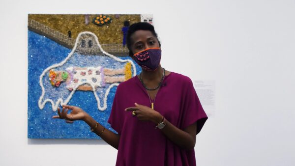 A black woman wearing a mask gestures to a painting behind her