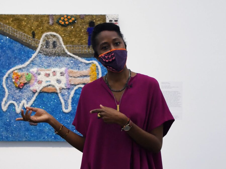 A black woman wearing a mask gestures to a painting behind her