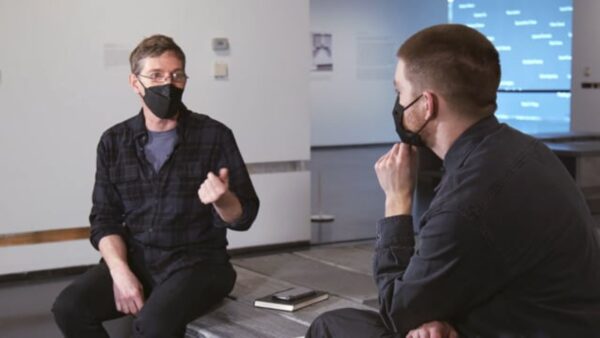 Two people sit in a gallery space wearing masks. They are positioned next to each other, but captured at an angle so only one person's face is visible.