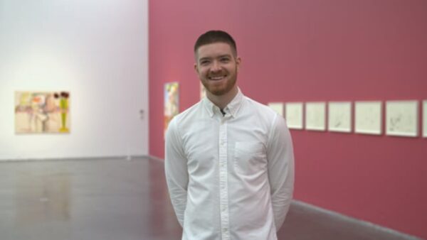 A male presenting person in a white button down smiles in a room with one wall painted pink, the adjoining wall painted white, with artworks hanging on each.