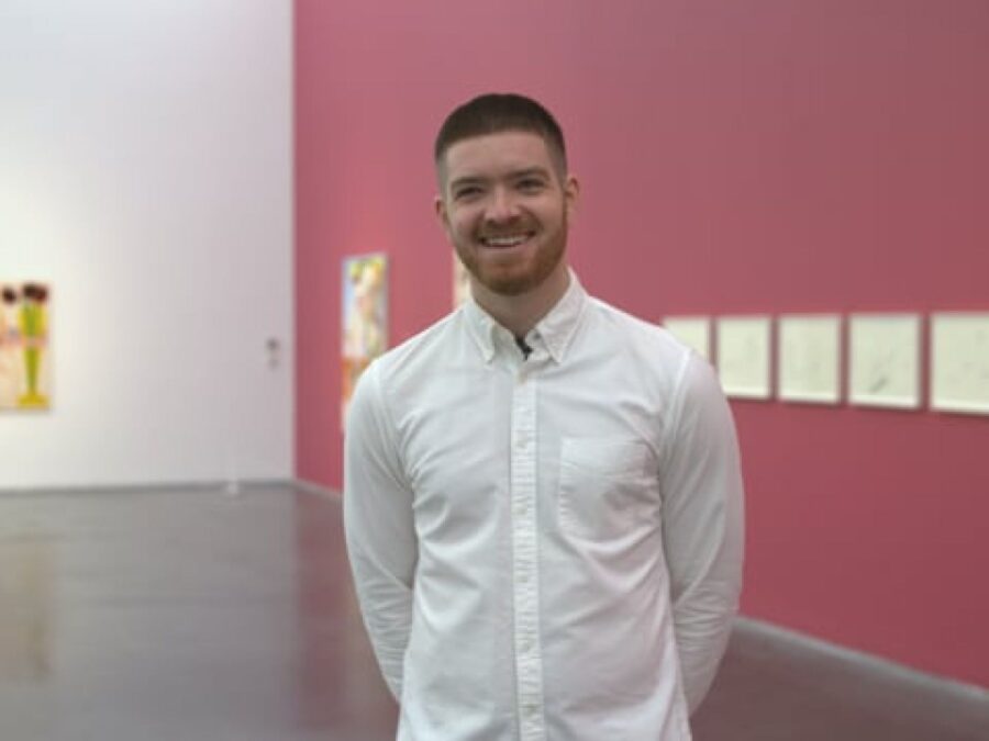 A male presenting person in a white button down smiles in a room with one wall painted pink, the adjoining wall painted white, with artworks hanging on each.