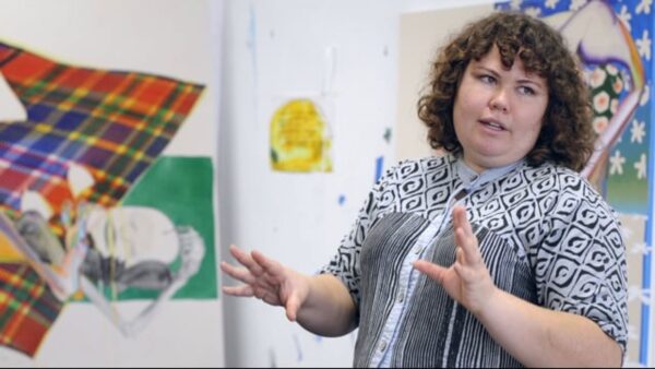 A female-presenting person with curly brown hair and pale ivory skin gestures with their hands in a white room filled with colorful artworks on the wall.