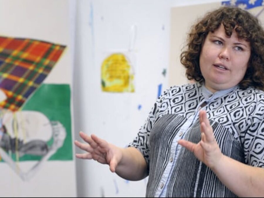 A female-presenting person with curly brown hair and pale ivory skin gestures with their hands in a white room filled with colorful artworks on the wall.