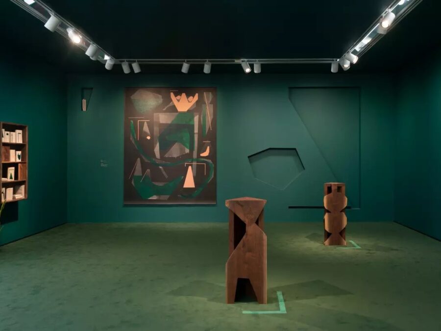 A room featuring teal-green walls and carpet with wooden sculptures on the floor and an abstract painting on the back wall