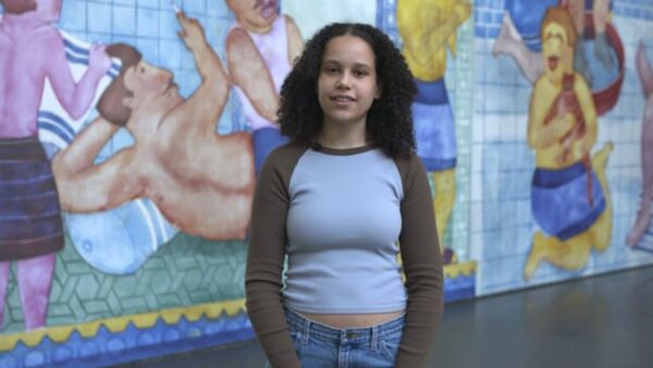 A female presenting person with brown curly hair and light tan skin stands against a large mural
