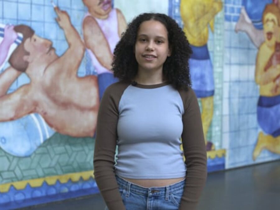 A female presenting person with brown curly hair and light tan skin stands against a large mural