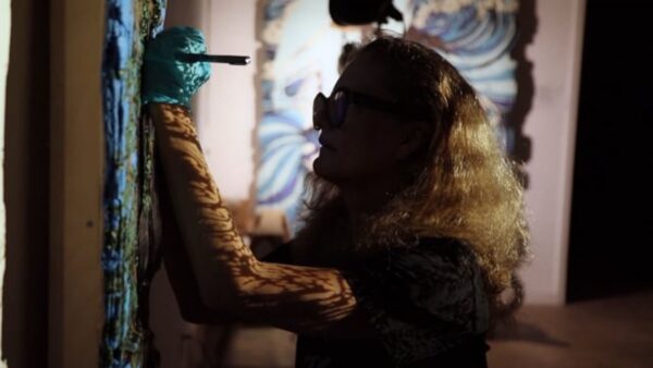 Dimly lit image of a person with full blond hair and glasses working on an artwork hanging on a wall.