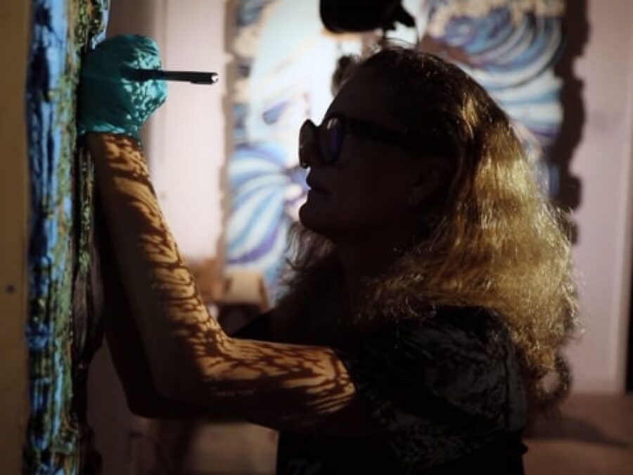 Dimly lit image of a person with full blond hair and glasses working on an artwork hanging on a wall.