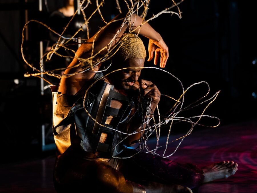 Jerron Herman, a dark-skinned Black man with blonde hair and a dark mustache, sits on the stage curled into a taut spiral; he is wrapped in a tangle of barbed wire. He wears a shimmery bodysuit and a black leather top. His eyes are closed, a tense expression on his face; one arm arcs over his head, palm open, as the other is tucked at his shoulder in a fist