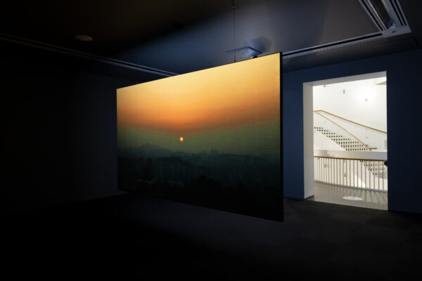 A film plays on a wall in a darkened room next to a doorway revealing a brightly lit staircase.