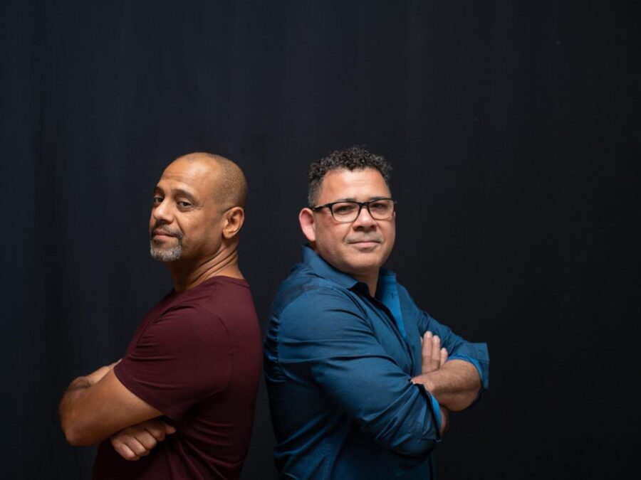 Two people pose back to back with their arms crossed in front of their bodies as they turn their heads toward the camera