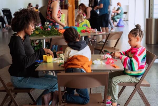 An adult and two children work on crafts at a table in a room with other participants