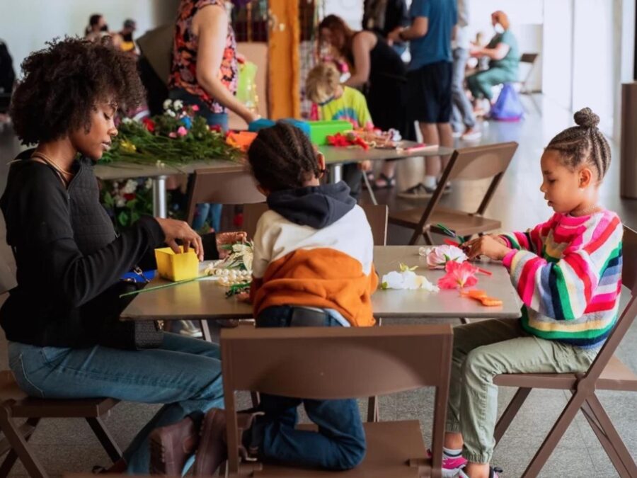 An adult and two children work on crafts at a table in a room with other participants