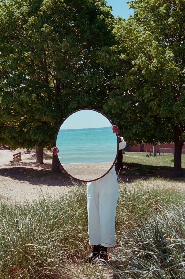 A person in the middle of a grassy area of a park setting holds a round mirror in front of their face which reflects an ocean setting.
