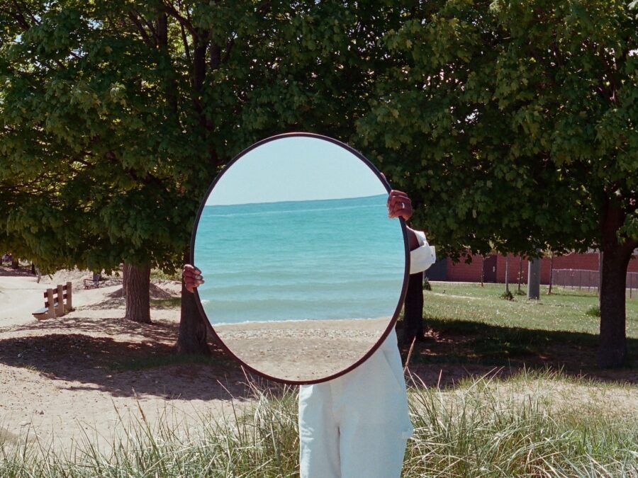 A person in the middle of a grassy area of a park setting holds a round mirror in front of their face which reflects an ocean setting.