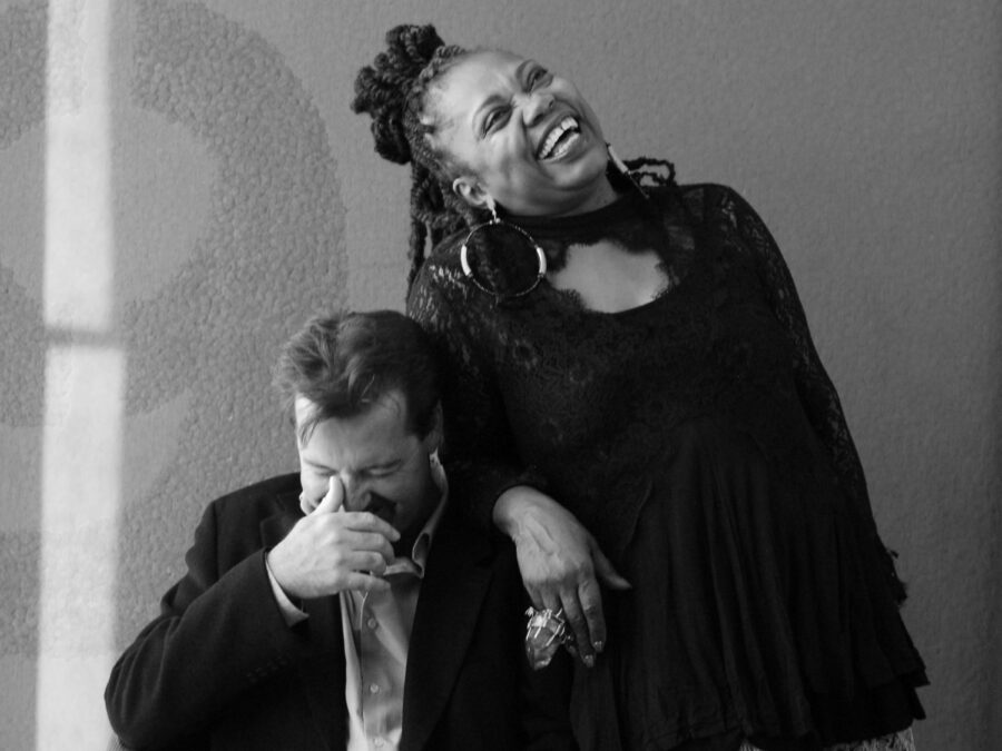 Black-and-white image of a man and a woman. The man is sitting down; the woman is standing, leaning on him. Both are laughing deeply.
