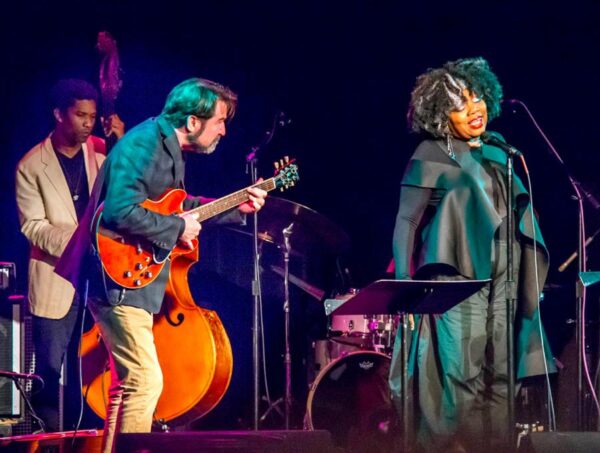 A guitarist takes cues from a singer on stage with additional band members in the background