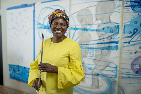 Art Professor Maria Magdalena Campos-Pons holding a long paintbrush in her studio office with colorful blue works behind her