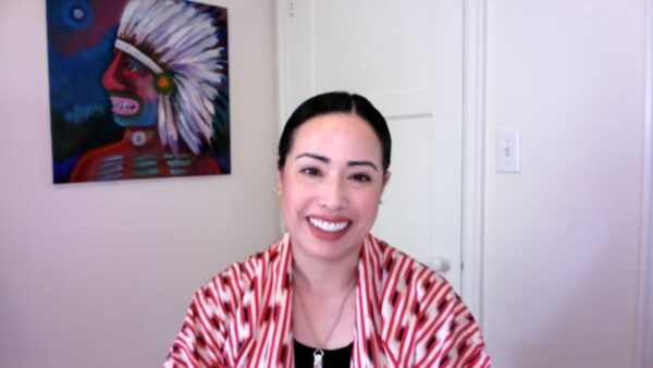A female presenting person with pale peach skin in a red and white patterned shirt smiles at the viewer from a room with a closed door behind them. On the wall is a painting of a person wearing a feather headdress.