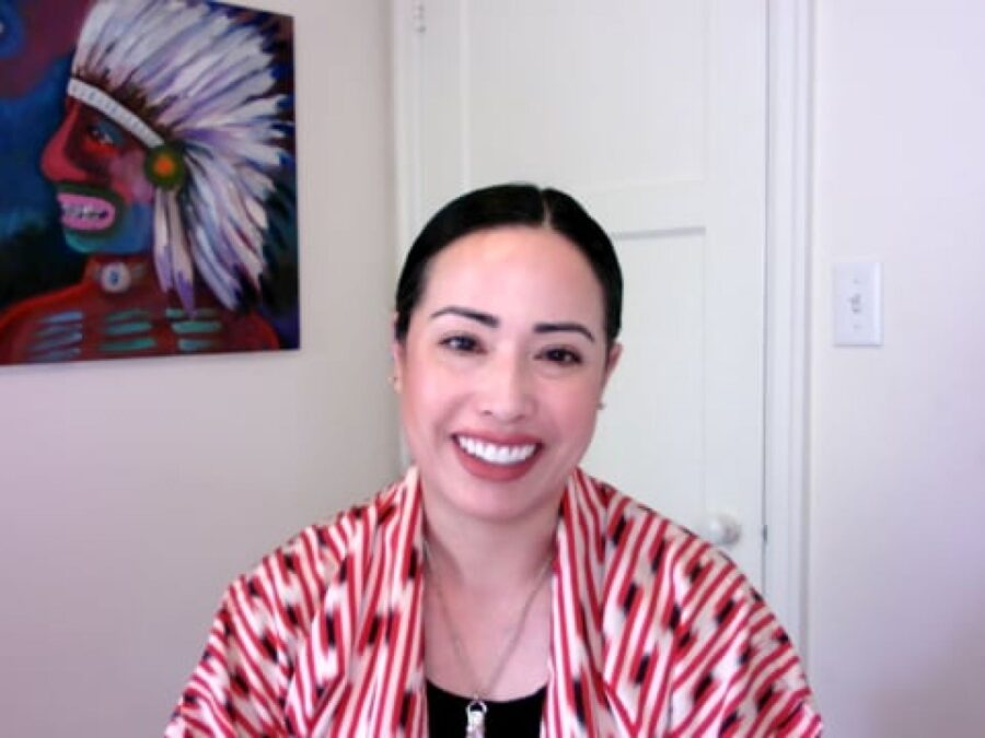 A female presenting person with pale peach skin in a red and white patterned shirt smiles at the viewer from a room with a closed door behind them. On the wall is a painting of a person wearing a feather headdress.