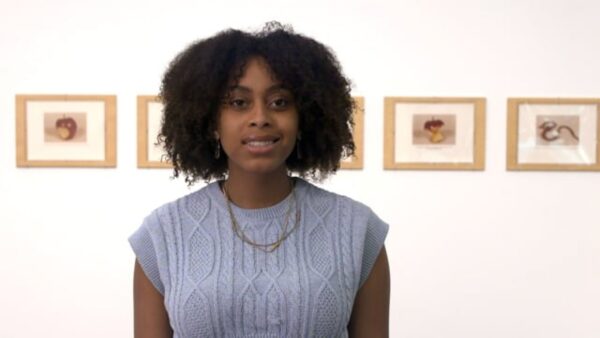 A female-presenting person with curly dark brown hair in front of a series of artworks on a wall
