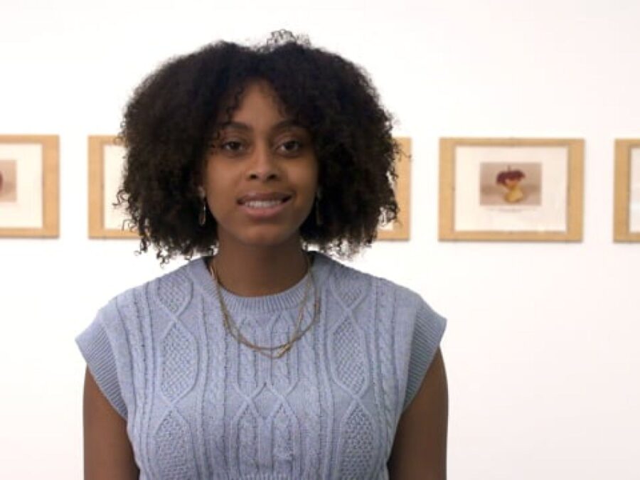 A female-presenting person with curly dark brown hair in front of a series of artworks on a wall
