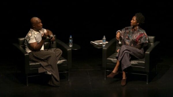 Two people sit kiddie corner from each other in chairs on a black stage