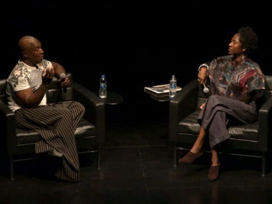 Two people sit kiddie corner from each other in chairs on a black stage
