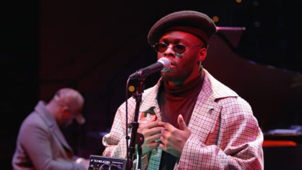 A male-presenting person with dark skin wearing sunglasses and a black fedora stands behind a microphone on a black stage lit by a red light