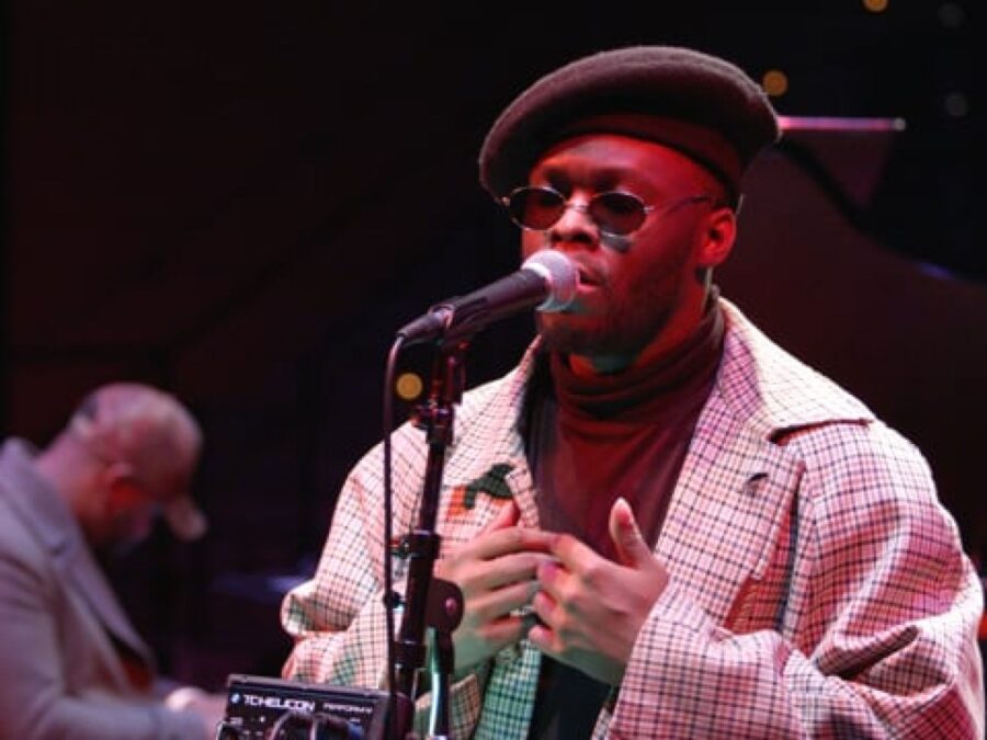 A male-presenting person with dark skin wearing sunglasses and a black fedora stands behind a microphone on a black stage lit by a red light