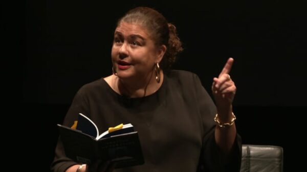 A female-presenting person with medium-dark skin holds a book in one hand while pointing with their other hand