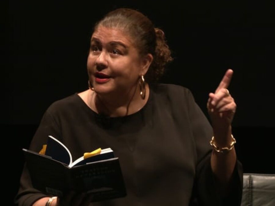 A female-presenting person with medium-dark skin holds a book in one hand while pointing with their other hand
