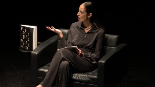 A female presenting person dressed in a black collared shirt and pants with dark hair pulled back gestures to someone off screen on a black stage. A book titled Forecast Form rests upright on a black table next to her.