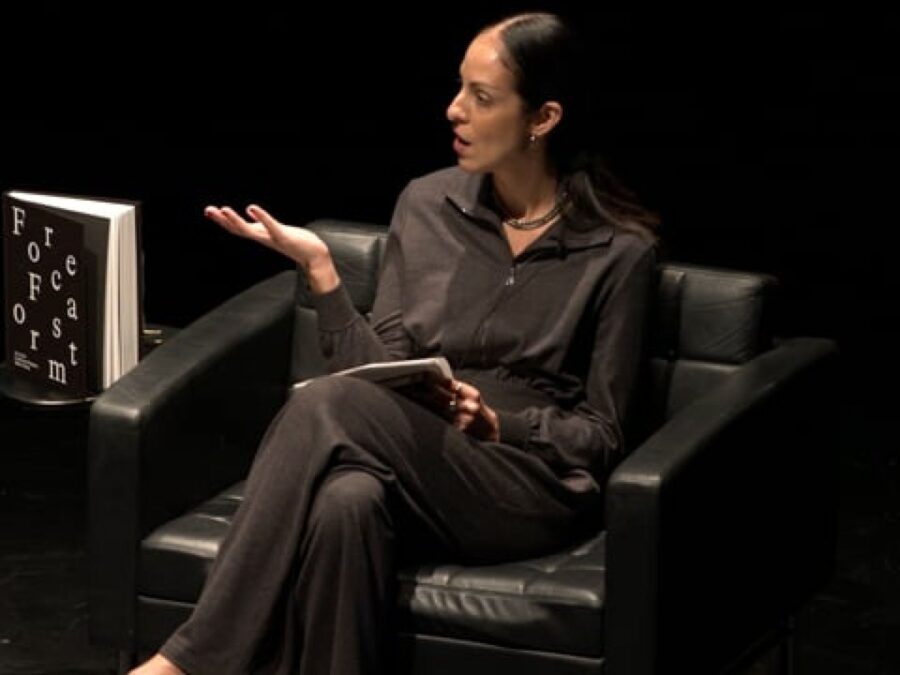 A female presenting person dressed in a black collared shirt and pants with dark hair pulled back gestures to someone off screen on a black stage. A book titled Forecast Form rests upright on a black table next to her.