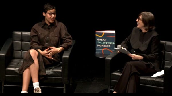 Two people sit on black chairs on a black stage with a book titled Great Woman Painters on a table between them