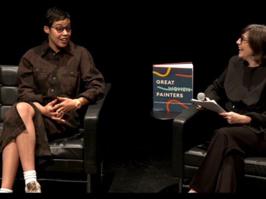 Two people sit on black chairs on a black stage with a book titled Great Woman Painters on a table between them