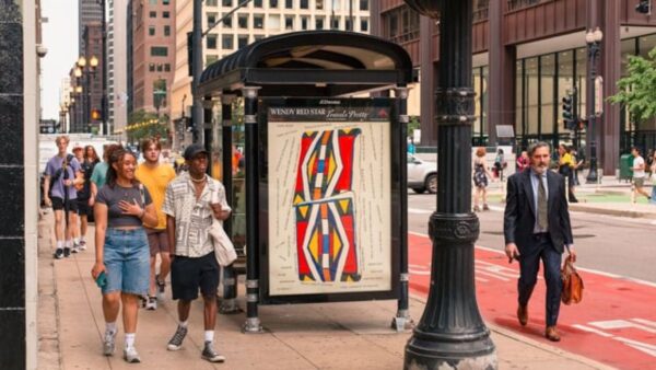 People walk past a bus shelter with a poster of a colorful, geometric Native artwork in the ad space