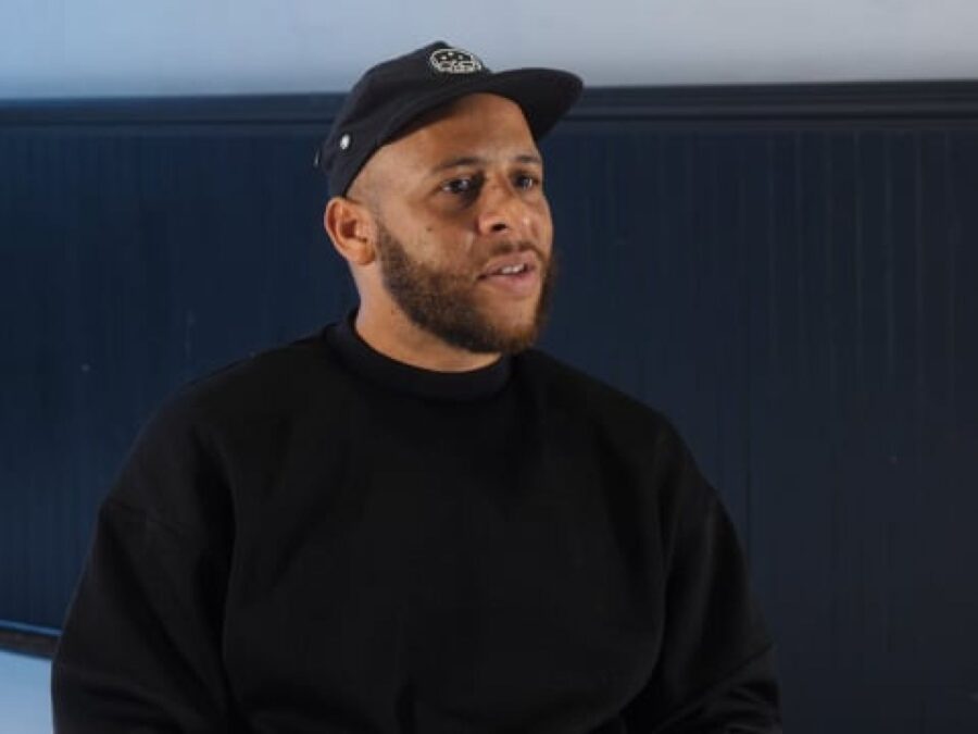 A male-presenting person from the waist up in a black sweatshirt and baseball cap with a close-cut beard in a room that has a long horizontal strip of gray on the wall