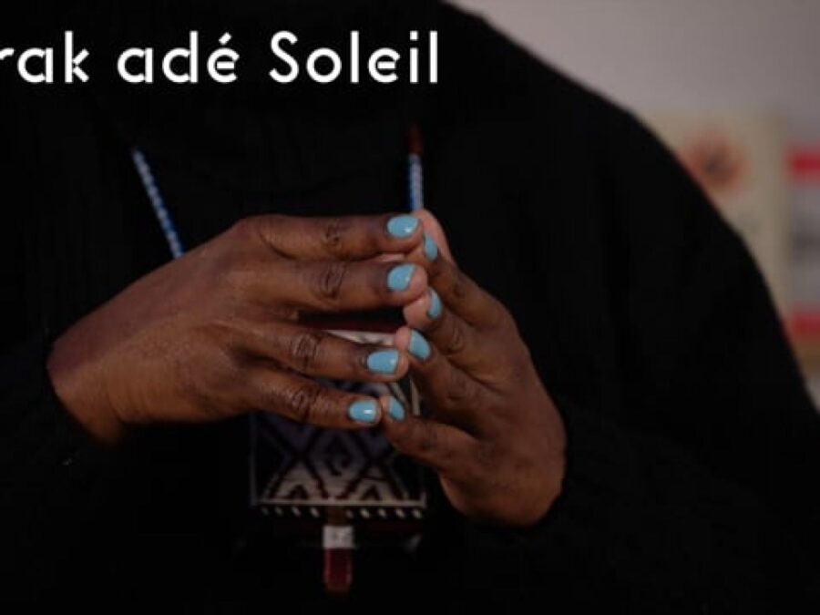 Close up of dark brown hands with robin's egg blue finger nails tented in front of somebody's chest wearing a black shirt. White text at the top of the image reads Barak ade Soleil.