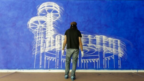 A person wearing a brown hat, black short-sleeved shirt, jeans, and white gloves stands with their back to the camera looking at an extra large painting of a white structure against a blue background