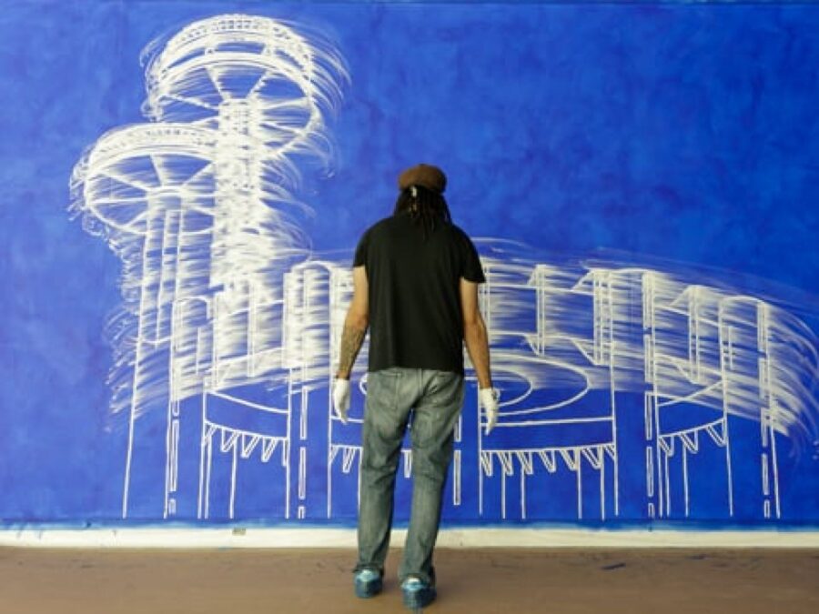 A person wearing a brown hat, black short-sleeved shirt, jeans, and white gloves stands with their back to the camera looking at an extra large painting of a white structure against a blue background