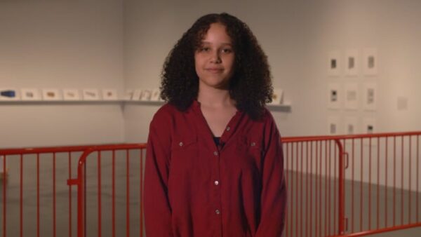 A teenager in front of a red railing looks directly at the camera