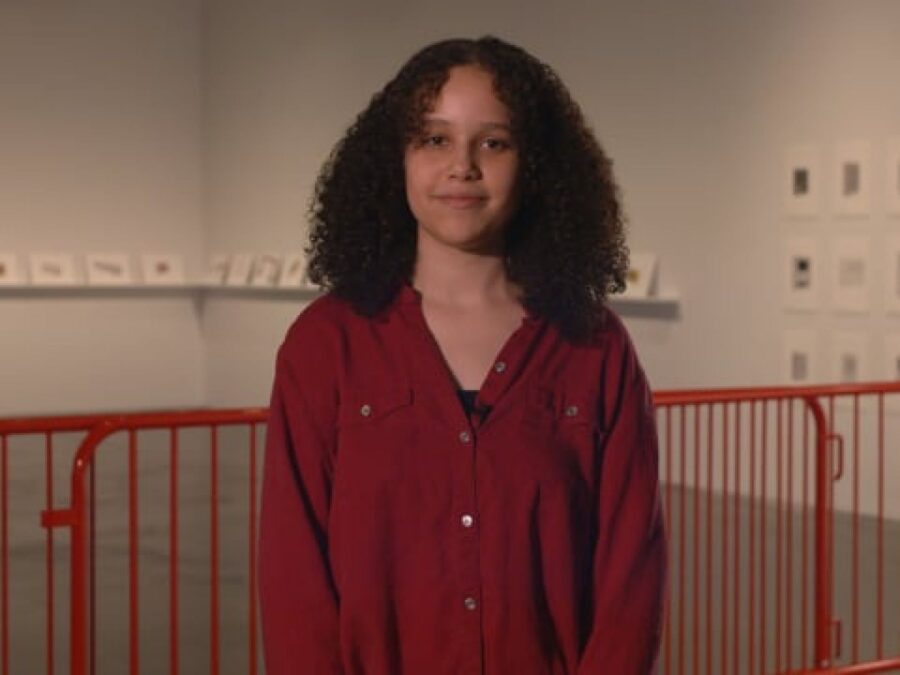 A teenager in front of a red railing looks directly at the camera