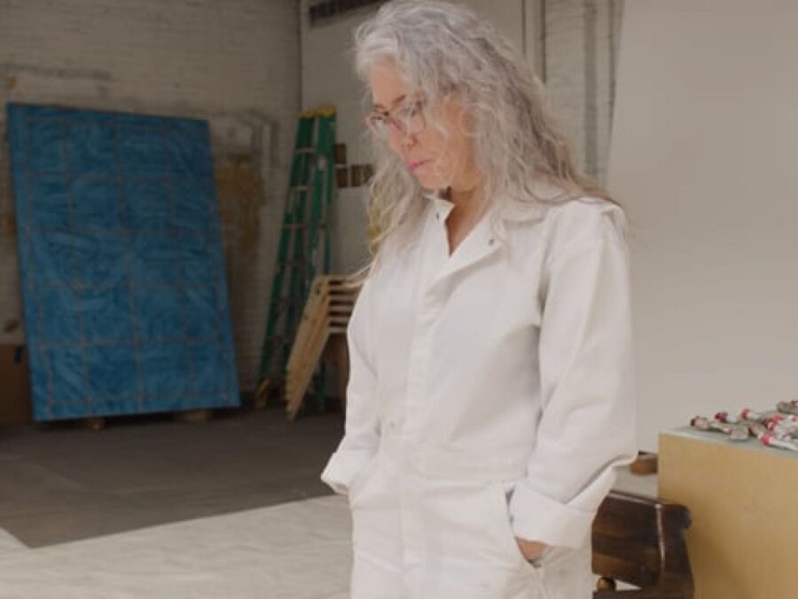 A female-presenting person with shoulder-length grey hair looks down at the floor in a studio space
