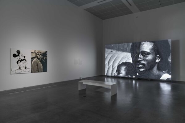 A screen displays a black-and-white image of a Black man and the top of a child's head in a darkened room with a white bench and an artwork featuring Mickey Mouse next to an image of a person with a painted white face.