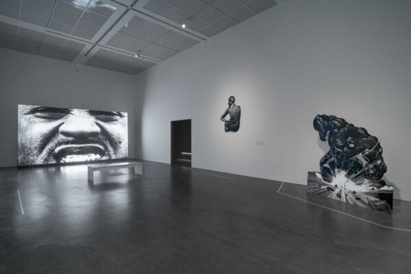 A screen displays a black-and-white close up of a person with a mustache and teeth exposed in a grin or grimace in a room with a bench and two other artworks.