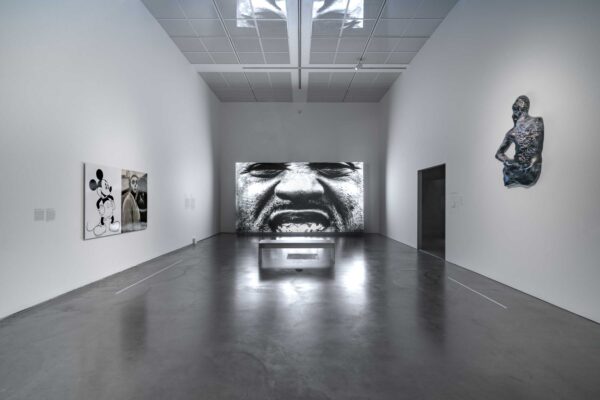 A dimly lit gallery with a screen displaying a black-and-white image of a person with a mustache grimacing and two other artworks on the two adjacent walls.